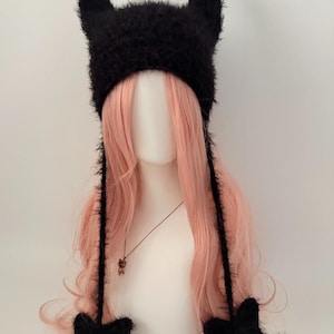 May include: A black, fuzzy hat with devil horns and star-shaped ear flaps. The hat is displayed on a mannequin head with long, pink hair. A gold necklace with a small pendant is also visible.
