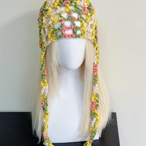 May include: A colorful crochet hat with a mesh design, featuring a mix of yellow, green, pink, and white yarn. The hat has long, braided ties that hang down on either side. The hat is displayed on a mannequin head with blonde hair.