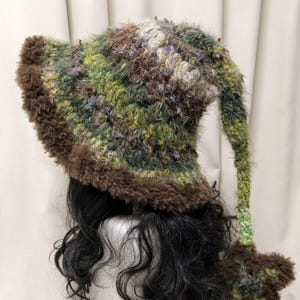 May include: A crochet hat featuring a variegated green, brown, and tan fuzzy yarn texture. The hat has a wide, dark brown faux fur brim and a long, dangling tail that ends in a dark brown star-shaped accent made of the same fuzzy material.