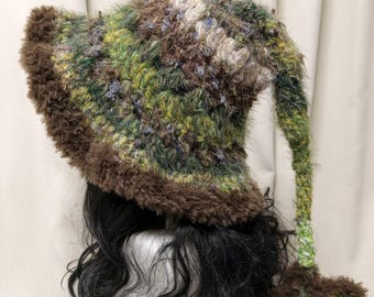 May include: A crochet hat featuring a variegated green, brown, and tan fuzzy yarn texture. The hat has a wide, dark brown faux fur brim and a long, dangling tail that ends in a dark brown star-shaped accent made of the same fuzzy material.