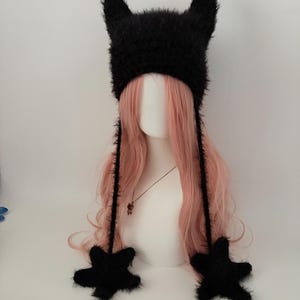 May include: A black, fuzzy hat with pointed ears and star-shaped accents. The hat has long, dangling ties and is displayed on a mannequin head with pink hair. The hat is a unique accessory.