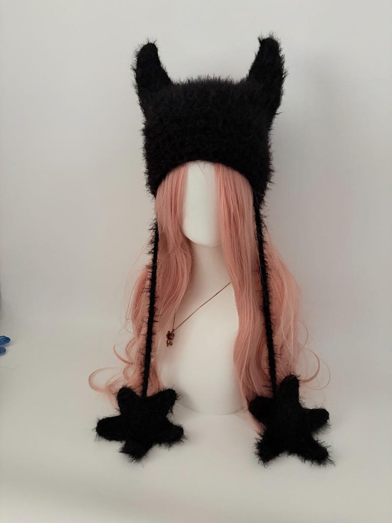 May include: A black, fuzzy hat with pointed ears and star-shaped accents. The hat has long, dangling ties and is displayed on a mannequin head with pink hair. The hat is a unique accessory.