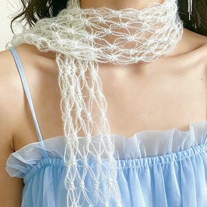 Peut inclure: Une &eacute;charpe blanche d&eacute;licate, &agrave; tissage ouvert, avec un design &agrave; franges. L'&eacute;charpe est drap&eacute;e autour du cou, mettant en valeur sa texture a&eacute;r&eacute;e et complexe. L'arri&egrave;re-plan pr&eacute;sente une robe bleu clair avec un col &agrave; volants.