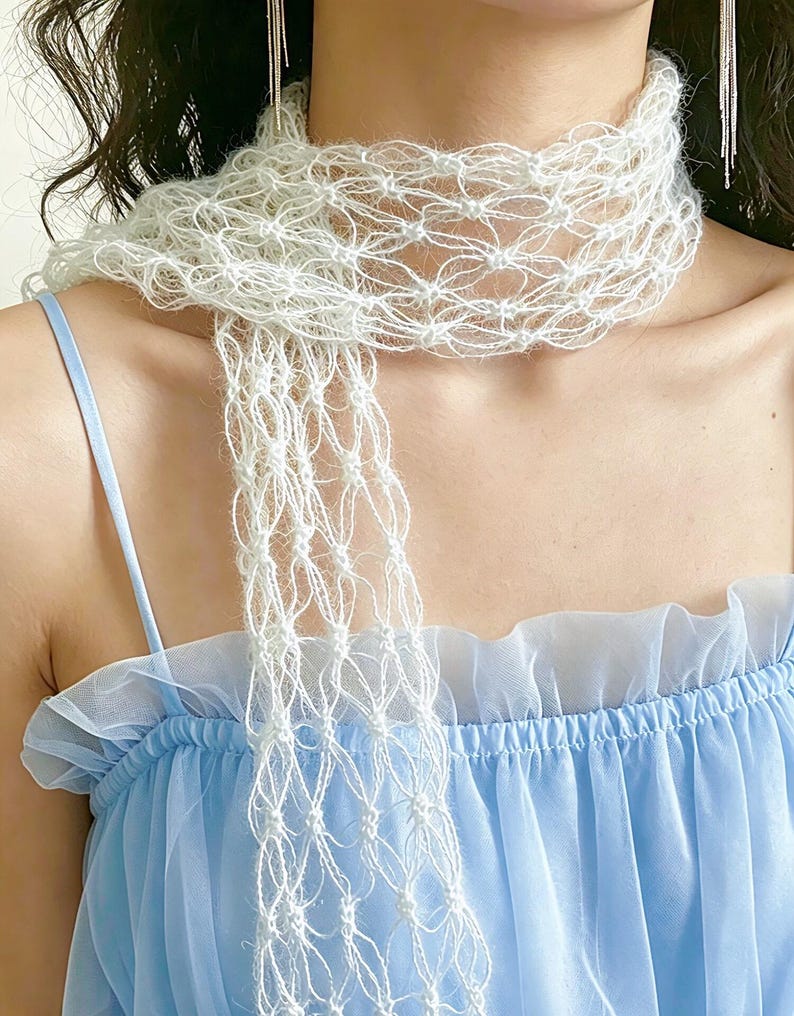 Peut inclure: Une &eacute;charpe blanche d&eacute;licate, &agrave; tissage ouvert, avec un design &agrave; franges. L'&eacute;charpe est drap&eacute;e autour du cou, mettant en valeur sa texture a&eacute;r&eacute;e et complexe. L'arri&egrave;re-plan pr&eacute;sente une robe bleu clair avec un col &agrave; volants.