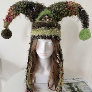 May include: A whimsical jester hat in shades of green, brown, and pink. The hat features three pointed tips with fluffy pom-poms and long, dangling strands. The textured fabric adds a unique, playful touch to this costume accessory.