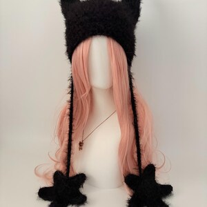 May include: A black fuzzy hat with two pointed horns and star-shaped ear flaps. The hat is displayed on a mannequin head with long, wavy pink hair. A necklace with a small pendant is visible.