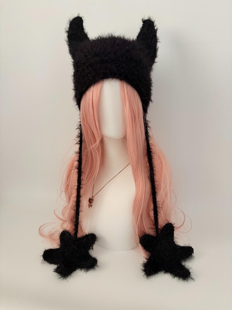 May include: A black fuzzy hat with two pointed horns and star-shaped ear flaps. The hat is displayed on a mannequin head with long, wavy pink hair. A necklace with a small pendant is visible.