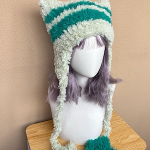 May include: A fuzzy, plush cat-ear beanie featuring horizontal stripes in alternating shades of mint green and deep teal. The hat includes two long, dangling earflaps that extend from the sides, each finished with a soft, textured pom-pom end.
