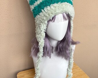 May include: A fuzzy, plush cat-ear beanie featuring horizontal stripes in alternating shades of mint green and deep teal. The hat includes two long, dangling earflaps that extend from the sides, each finished with a soft, textured pom-pom end.