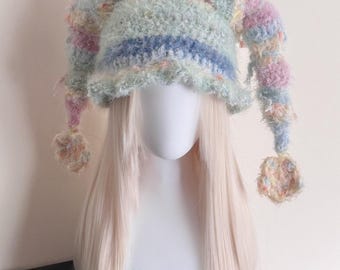 Peut inclure: Un chapeau de bouffon fait à la main aux couleurs pastel. Le chapeau présente un design duveteux et texturé avec des nuances de bleu, de rose, de jaune et de pêche. Il a de longues extrémités pendantes avec des pompons et est présenté sur une tête de mannequin avec de longs cheveux blonds.