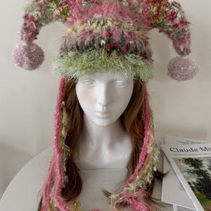 May include: A whimsical jester hat with pink, green, and white accents. The hat features two pointed tips with lavender pom-poms, a green band, and pink yarn braids. The hat is displayed on a mannequin head.