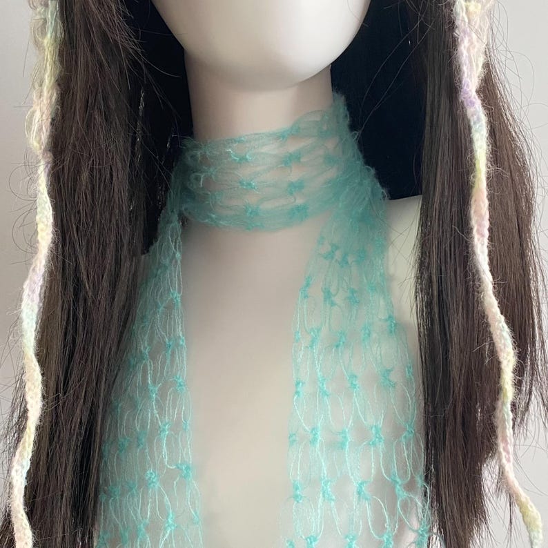 Peut inclure: Une &eacute;charpe bleu clair &agrave; tissage ouvert drap&eacute;e autour du cou d'un mannequin. L'&eacute;charpe a un aspect d&eacute;licat et a&eacute;r&eacute;. De longs cheveux bruns fonc&eacute;s avec des m&egrave;ches de fil de couleur pastel sont visibles de chaque c&ocirc;t&eacute; du visage du mannequin. L'&eacute;charpe est bleu pastel.