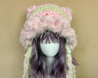 May include: A fuzzy pink bucket hat with cat-like ears, featuring a light green band and long, dangling braided fringe accents in shades of pink and lime green. The hat is adorned with small, colorful floral embellishments and beads.