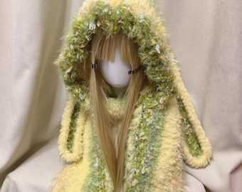 May include: A fuzzy, hooded scarf featuring long, dangling ear-like panels. The knit fabric is a blend of pale yellow, sage green, and white yarn with a shaggy, textured appearance. The hood is integrated into the scarf, creating a cohesive piece.