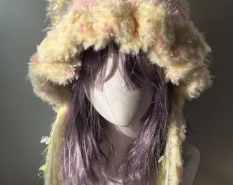 May include: A fuzzy, textured bonnet-style hat featuring a blend of pastel yellow, pink, and light green yarn. The hat has two pointed cat-ear shapes at the top and long, dangling side ties made of matching multi-colored, shaggy, and fringed yarn.