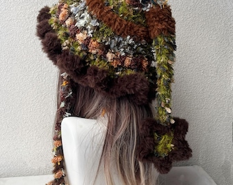 May include: A textured, multi-colored hat featuring a mix of brown, green, and orange yarn fibers. The hat has a wide, fuzzy brown brim, a long dangling side panel with a star-shaped end, and long braided tassels hanging down.