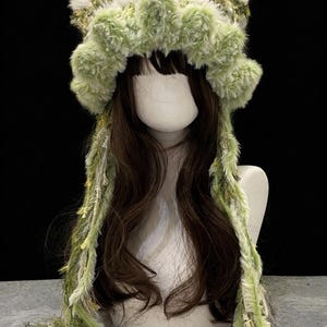 May include: A whimsical, green and white faux fur hat with long, dangling strands. The hat features a fluffy, textured design with floral accents. The hat is displayed on a mannequin head with long, brown hair, set against a dark background.