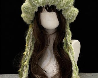 Peut inclure: Un chapeau fantaisiste en fausse fourrure verte et blanche avec de longues mèches pendantes. Le chapeau présente un design duveteux et texturé avec des accents floraux. Le chapeau est présenté sur une tête de mannequin avec de longs cheveux bruns, sur fond sombre.
