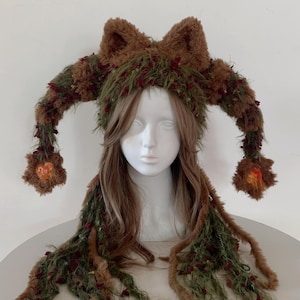 May include: A brown faux fur hat featuring two pointed ears and long, dangling side panels. The hat is decorated with textured green yarn and small red accents, with orange patches at the ends of the side panels and long fringe detailing.