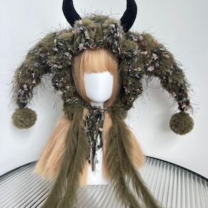 May include: A unique headpiece with an olive green and brown textured hood, complete with black horns. The hood is embellished with fluffy details, pink floral accents, and long, flowing olive green fringe. Two olive green pom-poms are attached to the sides.