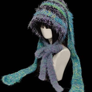 May include: A fuzzy, multi-coloured knit bonnet featuring long, dangling earflaps and a tie closure under the chin. The yarn is a textured blend of teal, purple, black, and lime green, creating a variegated appearance with a soft, shaggy finish.