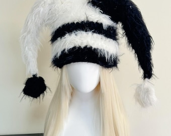 May include: A whimsical jester hat in black and white. The hat features alternating horizontal stripes and long, floppy, fur-like extensions. The hat is displayed on a mannequin head with long, blonde hair.