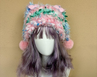 May include: A fuzzy, multi-colored knit beanie featuring a blend of pink, light blue, and green yarn. The hat has two small, rounded ear-like protrusions at the top and two pink pom-poms hanging from the sides, resting on a mannequin head.