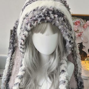 May include: A plush, hooded hat featuring long, dangling ear-like flaps. The exterior is crafted from a textured, faux-fur material in a mottled pattern of white, grey, and soft pink, with a solid white band running across the top of the hood.