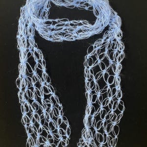 Peut inclure: Une &eacute;charpe bleu clair &agrave; tissage ouvert. L'&eacute;charpe est faite d'une mati&egrave;re douce et duveteuse et a un aspect d&eacute;licat et a&eacute;r&eacute;. Le motif pr&eacute;sente un motif r&eacute;p&eacute;titif de petites boucles interconnect&eacute;es, cr&eacute;ant un effet de dentelle. L'&eacute;charpe est pr&eacute;sent&eacute;e sur un fond sombre.