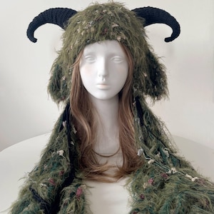 May include: A green fuzzy hat featuring two large black crocheted horns extending from the sides. The hat has long, shaggy ear flaps that drape down, incorporating textured green yarn with small flecks of pink and white throughout the material.