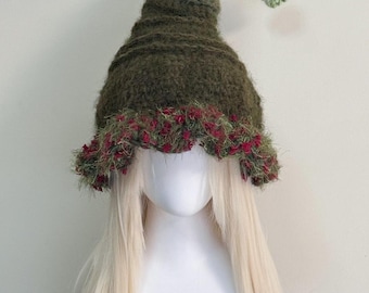 May include: A whimsical, handmade olive green crochet hat with a pointed crown and a decorative brim. The brim is adorned with red and green fuzzy yarn. The hat has a curved, green and white crocheted tip with a small green flower.