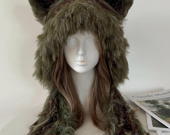 May include: A fuzzy olive green faux fur hat featuring prominent pointed animal ears on top. The hat has long, dangling side panels with a textured, shaggy appearance, incorporating subtle dark brown and maroon fibers throughout the material.