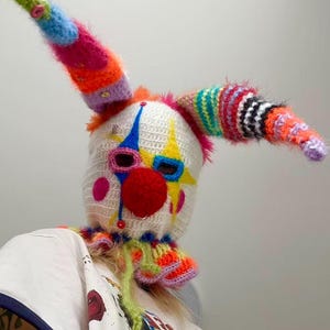 May include: A handmade, colorful clown mask with a white base, featuring a red nose, blue and yellow accents, and long, striped horns. The mask is crocheted with vibrant yarns and has a playful, whimsical design.