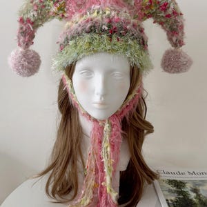Puede incluir: Un sombrero de bufón hecho a mano en tonos rosa, verde y amarillo. El sombrero presenta un diseño esponjoso y texturizado con tres pompones redondeados y una larga bufanda fluida. El sombrero se exhibe en una cabeza de maniquí.