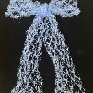 Peut inclure: Un n&oelig;ud papillon fait &agrave; la main, bleu clair, avec de longues queues fluides. Le n&oelig;ud est fait d'un fil d&eacute;licat &agrave; tissage ouvert, cr&eacute;ant un effet de dentelle. Le n&oelig;ud est nou&eacute; au centre avec un n&oelig;ud, et les queues pendent. Le fond est noir.
