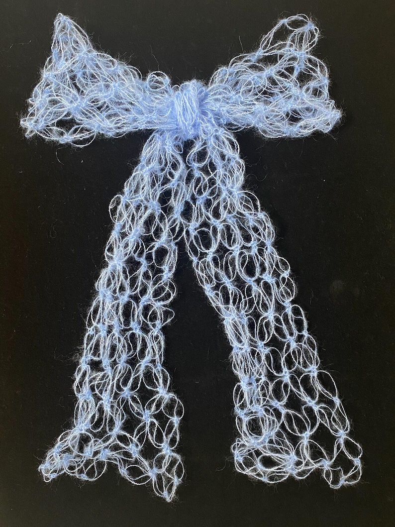 Peut inclure: Un n&oelig;ud papillon fait &agrave; la main, bleu clair, avec de longues queues fluides. Le n&oelig;ud est fait d'un fil d&eacute;licat &agrave; tissage ouvert, cr&eacute;ant un effet de dentelle. Le n&oelig;ud est nou&eacute; au centre avec un n&oelig;ud, et les queues pendent. Le fond est noir.
