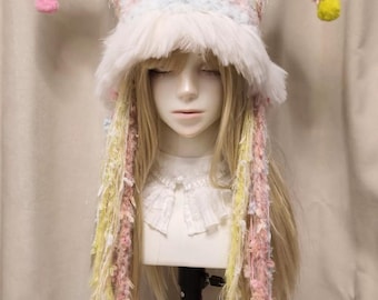 May include: A jester-style hat featuring two long, pointed horns with pom-poms at the ends. The hat is crafted from a multi-colored, textured yarn in pastel shades of pink, yellow, and blue, with a thick white faux fur trim along the brim.