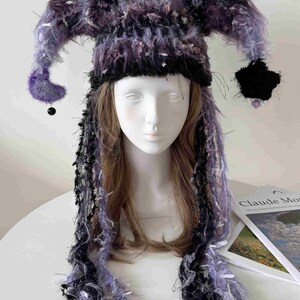 May include: A jester-style hat crafted from fuzzy, textured yarn in shades of purple and black. The hat features two long, drooping side points with small black beads at the ends and long, dangling fringe strands that hang down past the shoulders.
