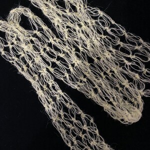 Peut inclure: Textile cr&egrave;me &agrave; tissage ouvert avec un motif en forme de filet. Le mat&eacute;riau semble &ecirc;tre fait d'un fil doux et fibreux, peut-&ecirc;tre de la laine ou une fibre naturelle similaire. Le textile est pr&eacute;sent&eacute; sur un fond noir, soulignant sa texture et son design.