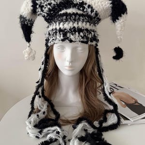 May include: A black and white jester hat with long, fringed trim. The hat features a striped pattern and dangling star and crescent-shaped accents. The hat is made of a soft, textured material.
