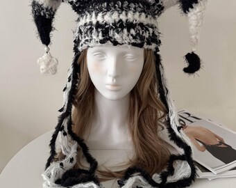 May include: A black and white jester hat with long, fringed trim. The hat features a striped pattern and dangling star and crescent-shaped accents. The hat is made of a soft, textured material.