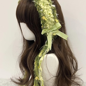 May include: A green and yellow headband with a textured, fuzzy design. The headband features a gingham bow and a long, decorative strand with small floral accents. The accessory is displayed on a mannequin head with brown hair.