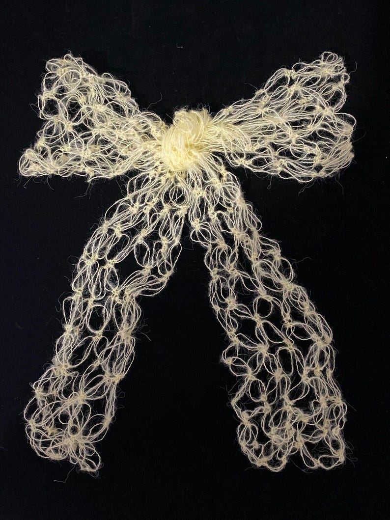 Peut inclure: Un n&oelig;ud papillon fait &agrave; la main de couleur cr&egrave;me, r&eacute;alis&eacute; en fil nou&eacute;. Le n&oelig;ud pr&eacute;sente un n&oelig;ud central et deux longues boucles fluides. Le fil a un aspect l&eacute;g&egrave;rement textur&eacute;, sur fond noir.