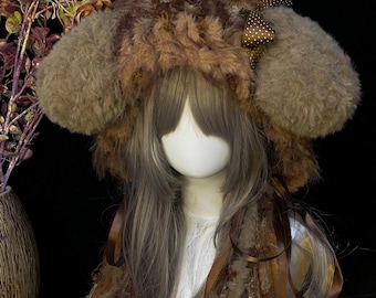 May include: A brown faux fur bonnet featuring large, rounded ear-like side panels, a brown polka-dot ribbon bow on the side, and long, textured hanging ribbons and fabric strips in shades of brown and tan, worn over a light-colored wig.