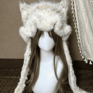 Può includere: Un cappello in pelliccia sintetica bianca con orecchie da gatto prominenti e lunghi pannelli laterali pendenti. Il cappello è realizzato con un materiale testurizzato e ispido e include elementi decorativi a frange lungo i pannelli laterali.