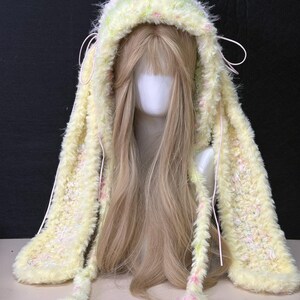May include: A fuzzy, pale yellow hooded accessory with long, dangling ear-like panels. The knit texture features subtle flecks of pink and white throughout. Thin, light pink cords are attached to the sides, hanging down alongside the long panels.