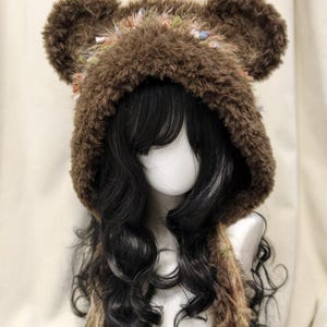 May include: A brown faux fur hooded hat featuring two rounded bear-like ears on top. The hood is lined with a textured, multi-coloured fringe trim in shades of pink, green, and blue. Long, matching brown faux fur ties hang down from the sides.