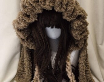May include: A brown faux fur hood with a textured, ruffled trim and long side panels. The hood features a lighter beige, fuzzy interior lining and two long, matching braided ties hanging down from the front, showcasing a soft, plush appearance.