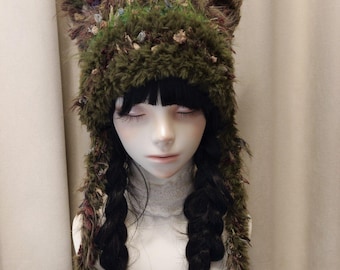 May include: A fuzzy olive green knit hat featuring two pointed cat-like ears at the top and long, dangling side panels. The yarn has a textured, multi-colored appearance with flecks of brown, tan, and muted red woven throughout the material.