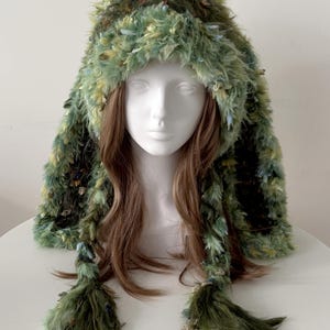 Puede incluir: Un sombrero de piel sintética verde y amarillo con capucha y largas borlas trenzadas. El sombrero está hecho de un material peludo y texturizado y se muestra en una cabeza de maniquí blanca. El sombrero tiene una apariencia caprichosa y de bosque.